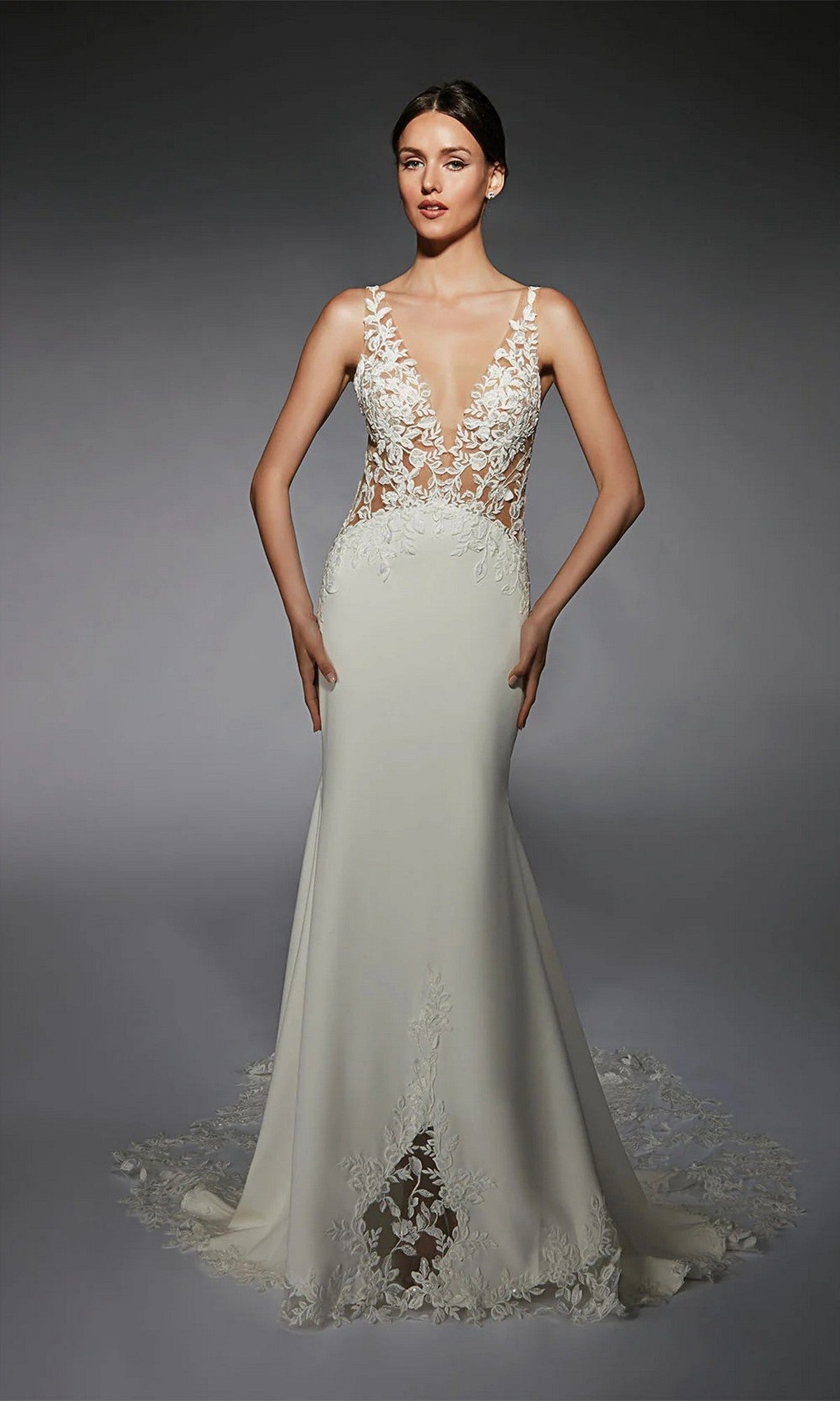 A woman poses against a dark background wearing the Alyce 7117 long white dress in ivory/tan, featuring a sheer lace bodice, deep V neckline, and lace detailing on the skirt and train.